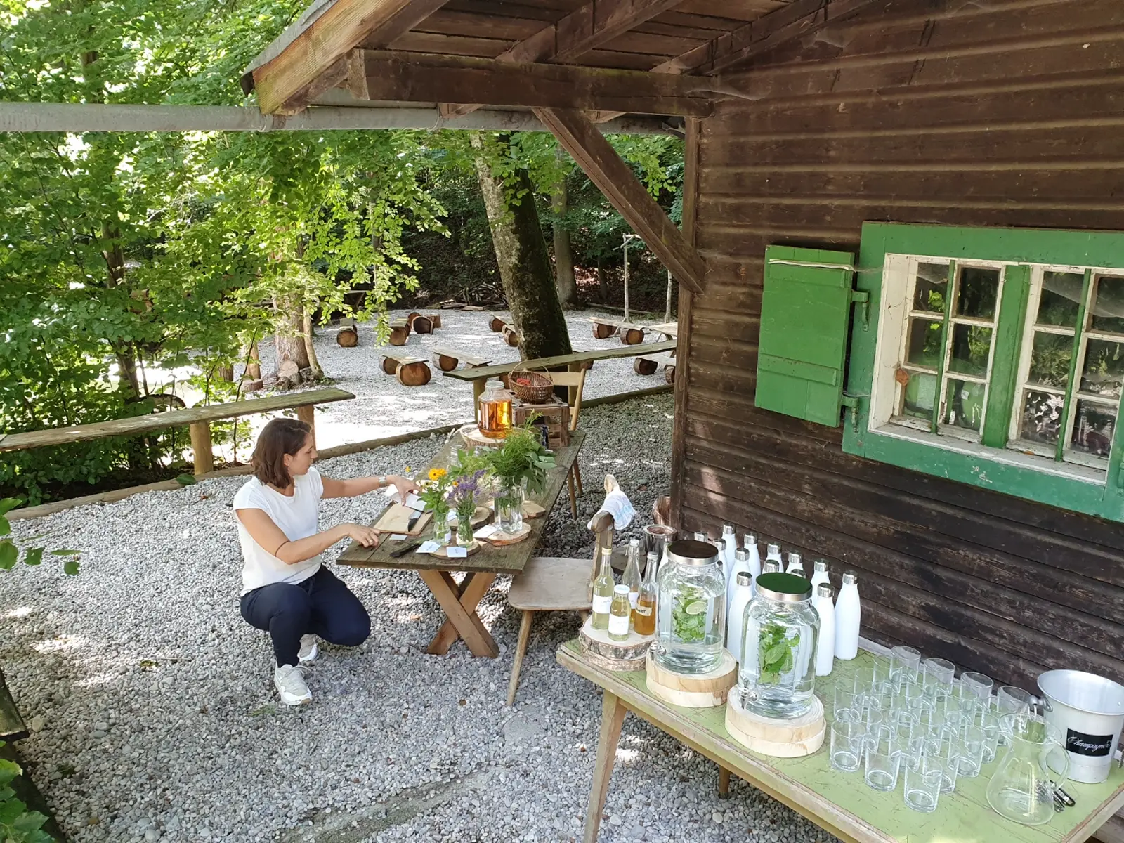 Workshop-Aufbau am Hüttenvordach — Holztisch mit Wildblumen, Glaskaraffen und Bar-Tafel vor dem Hüttenfenster